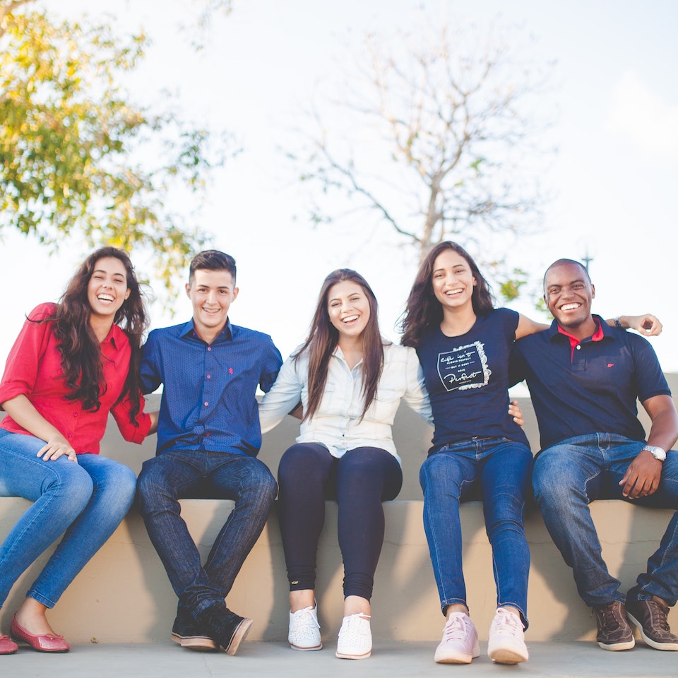 A group of ethnically diverse young women and men wearing jeans and red and blue tops, smiling, and sitting on a wall